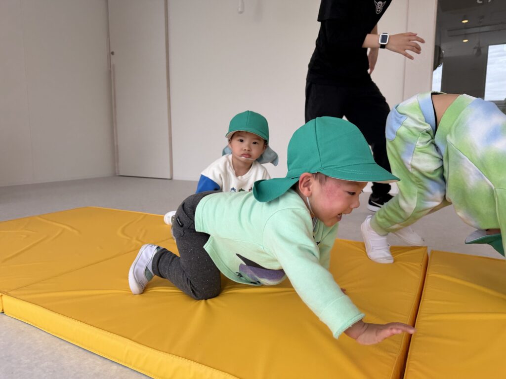 Two young children in green hats crawl on a yellow gym mat while a supervising adult stands nearby.